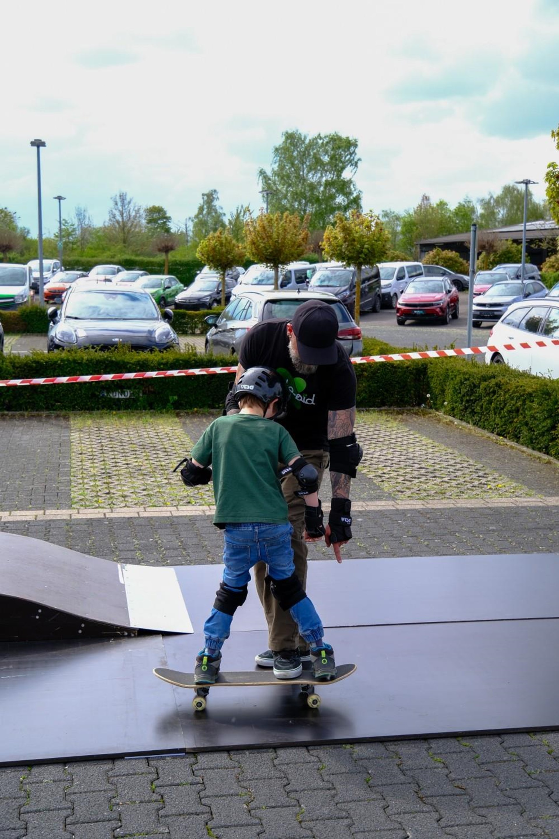 Auf dem Foto sieht man einen Jungen mit Helm und Knieschützern auf einem Skateboard. Ein Mann steht ihm Gegenüber und erklärt ihm etwas. 
Im Hintergrund ist ein Parkplatz zu sehen