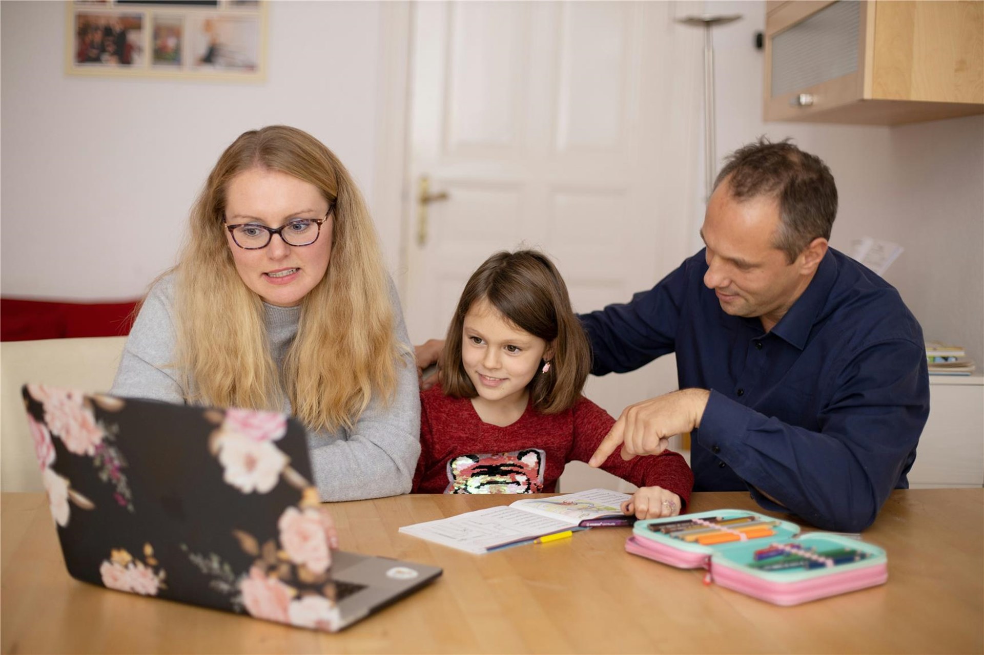 Ein Elternpaar sitzt zuhause am Tisch mit ihrer Tochter. Alle drei schauen auf einen Laptop. 