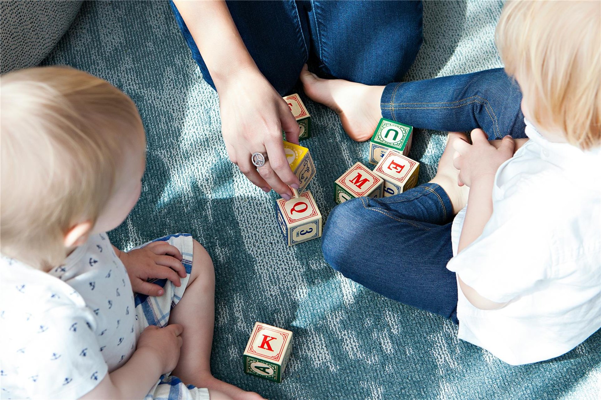Das Foto zeigt von oben, wie eine Frau und zwei Kinder auf einer Decke sitzen und mit Holzwürfeln spielen, auf denen Buchstaben abgebildet sind. 