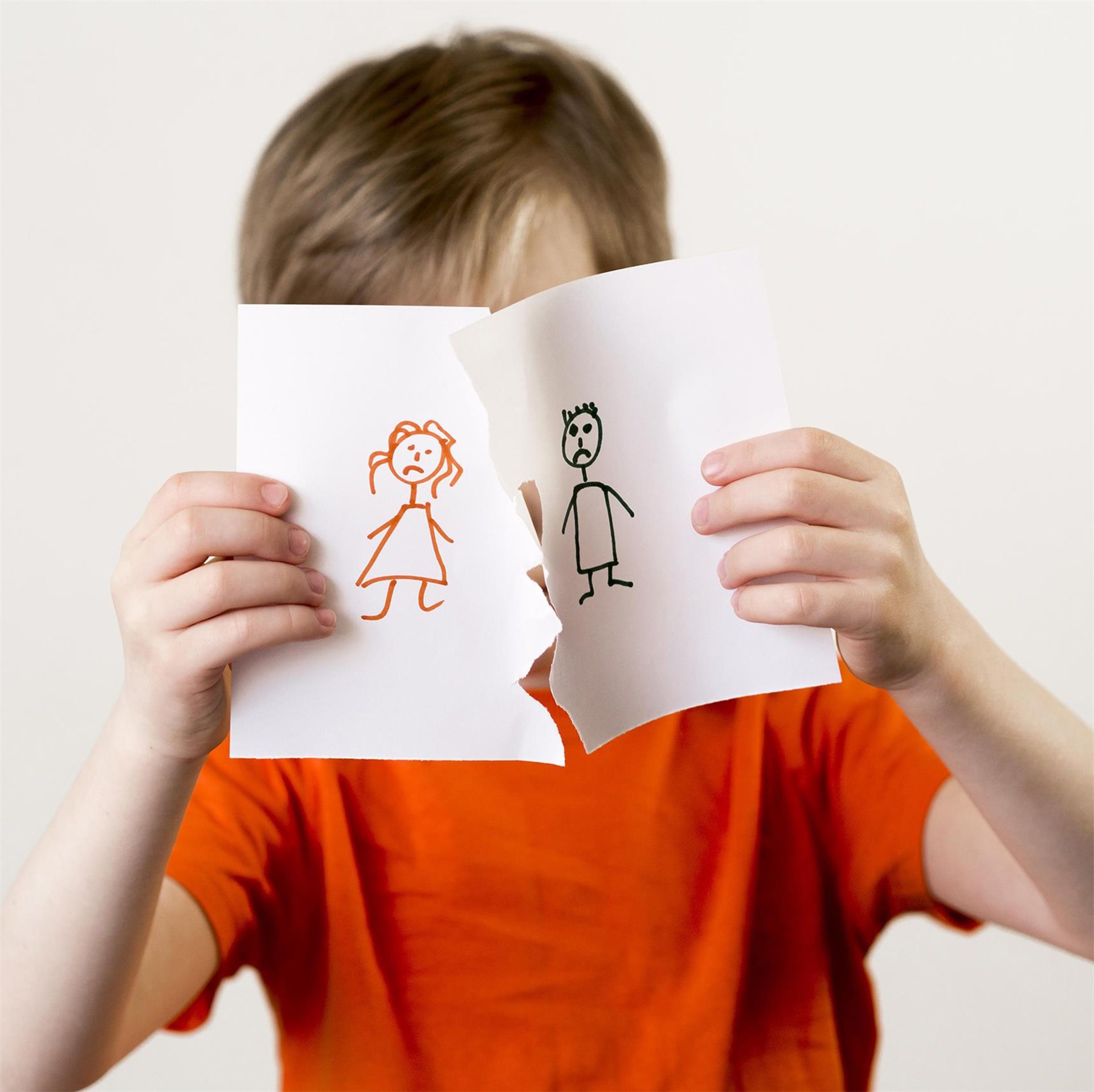 Ein Junge in einem orangefarbenen T-Shirt halt ein Bild mit Strichmännchenfiguren, die ein Ehepaar darstellen vor sich. Das Bild hat unten einen Riss. 