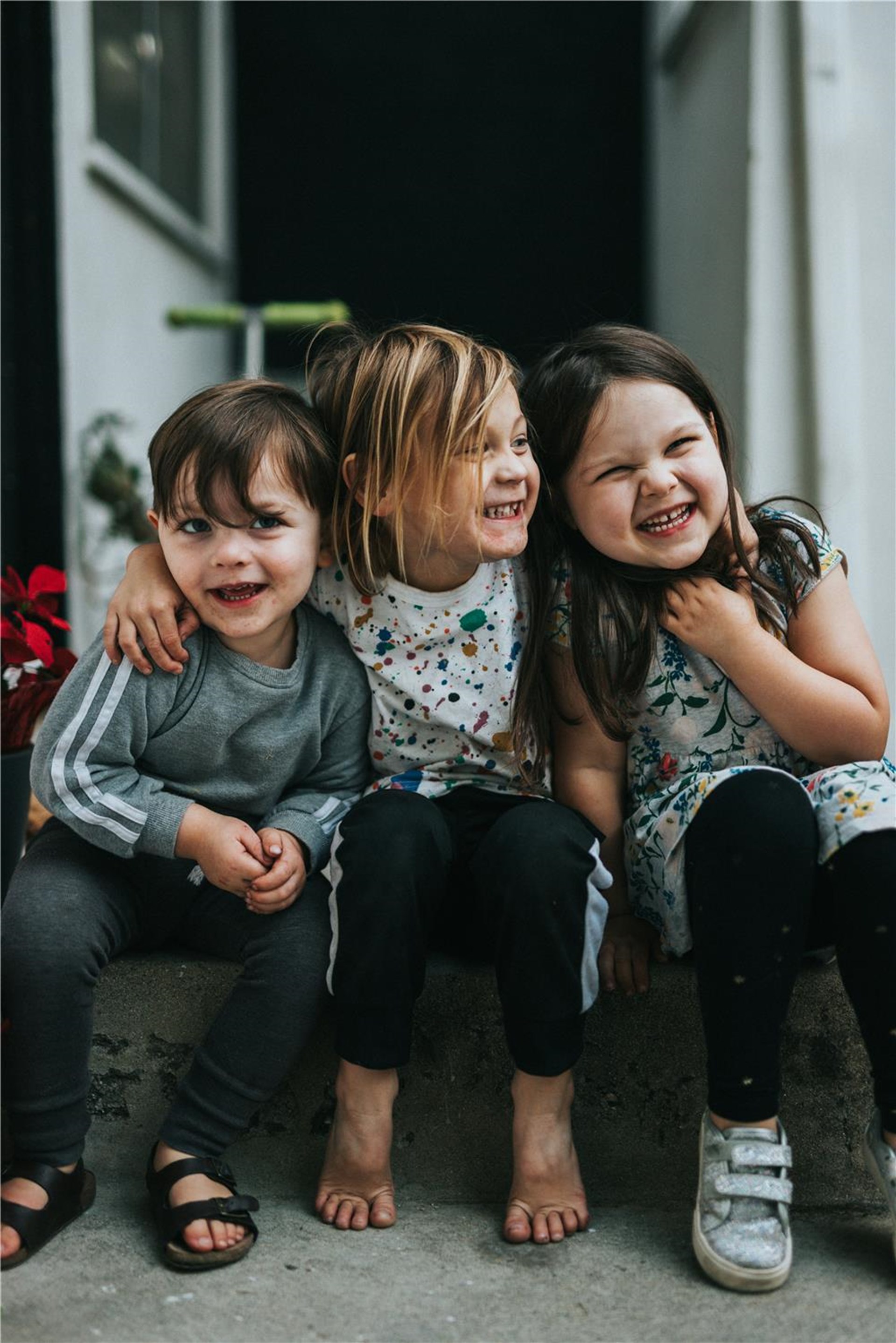 Drei Kinder sitzen zusammen auf einer Treppe. 
Das Mädchen in der Mitte legt ihre Arme um die beiden äußeren Kinder. 
