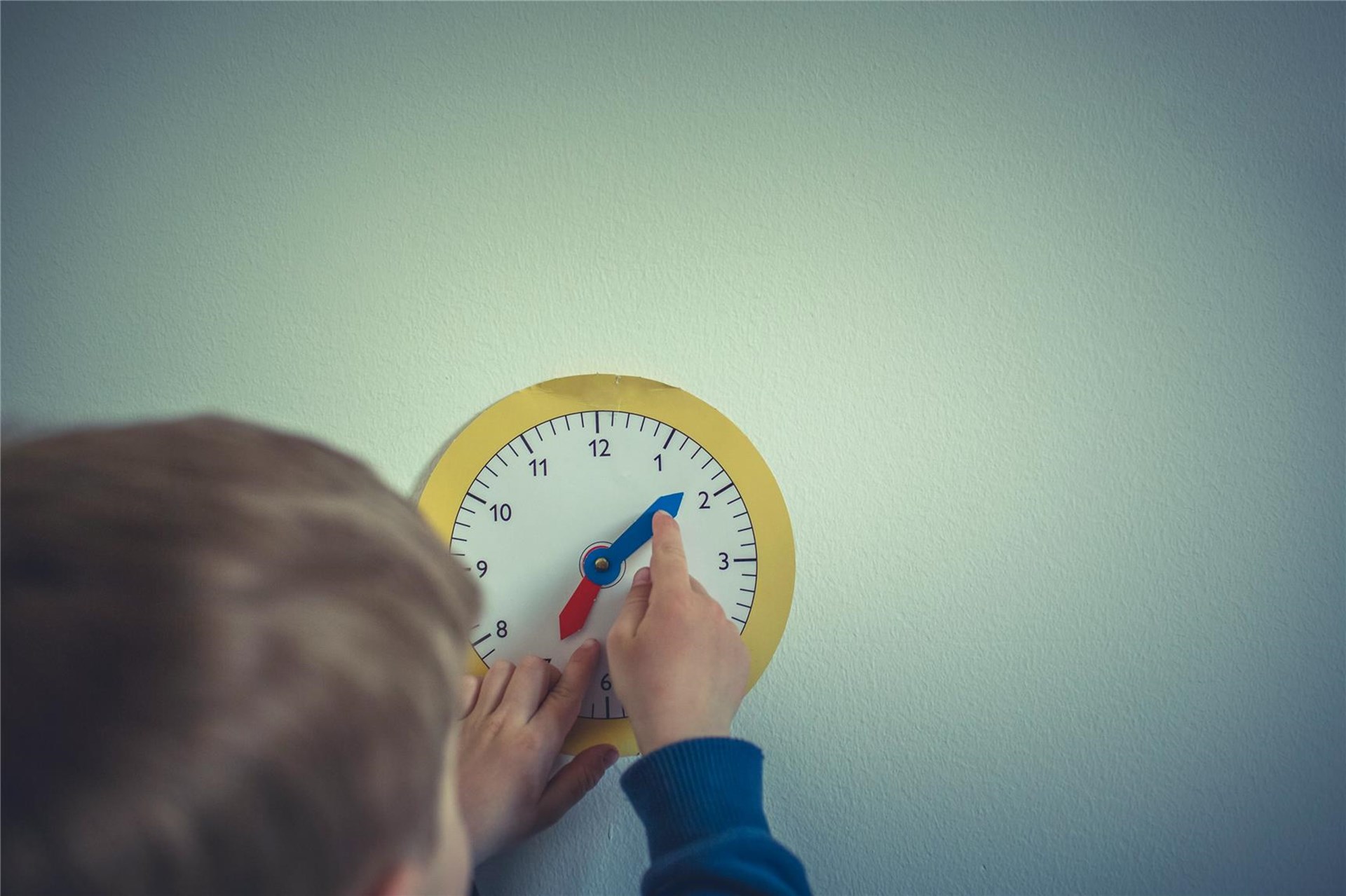 Ein Junge stellt den Zeiger einer Uhr ein. Er ist von hinten abgebildet. 
Man sieht nur den Hinterkopf und die Hand. 