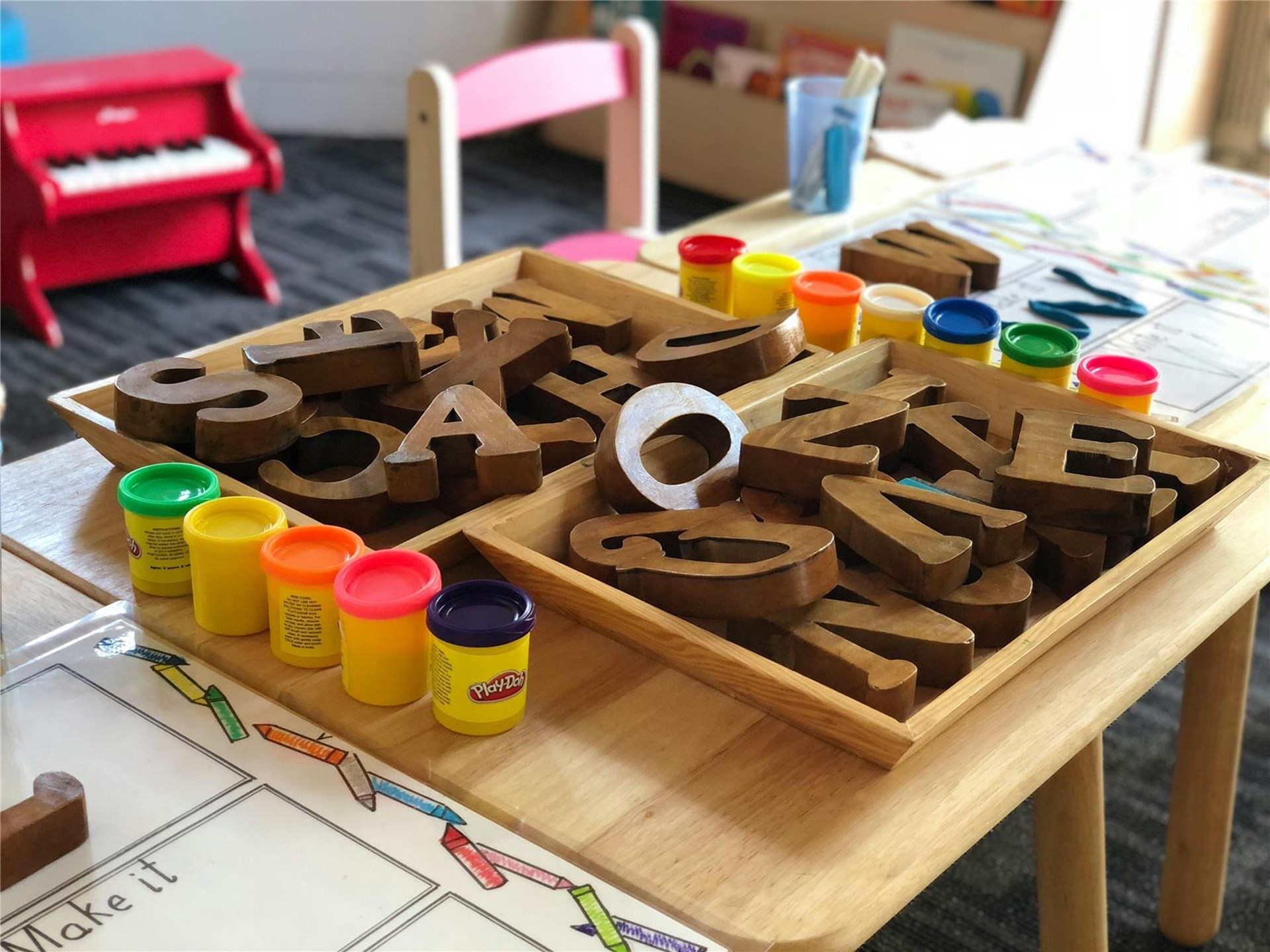 Das Bild zeigt einen Tisch im Kindergarten, auf dem Spielmaterialien liegen: Holzbuchstaben und Farbtöpfe