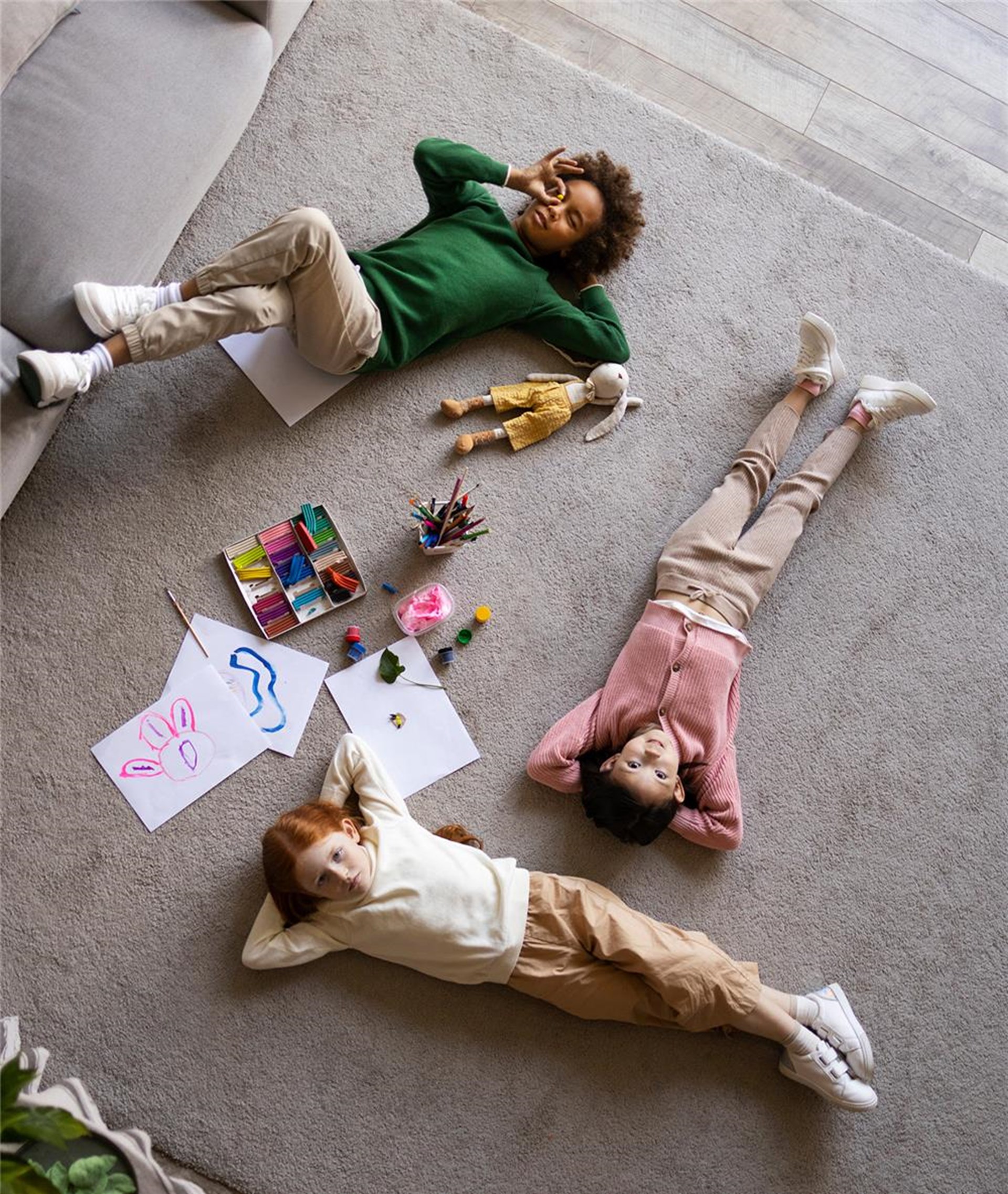 Drei Kinder liegen auf dem Rücken auf einem grauen Teppich. In ihrer Mitte sind Malsachen. Das Foto ist von oben aufgenommen. 